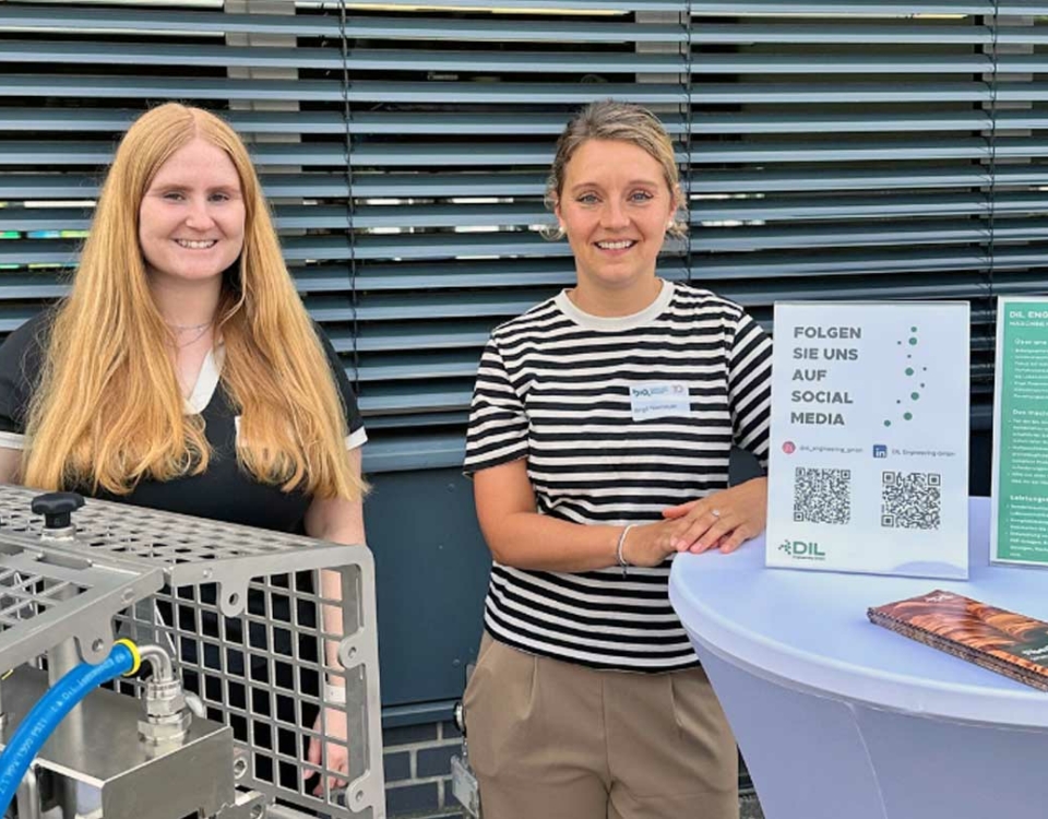 Zwei lächelnde Frauen an einem Informationsstand mit technischem Exponat, Social-Media-Schild und Werbematerialien.